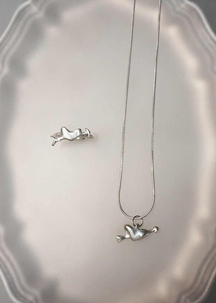 A silver necklace with a small abstract heart pendant, displayed on a scalloped white surface alongside a matching small silver accessory piece.