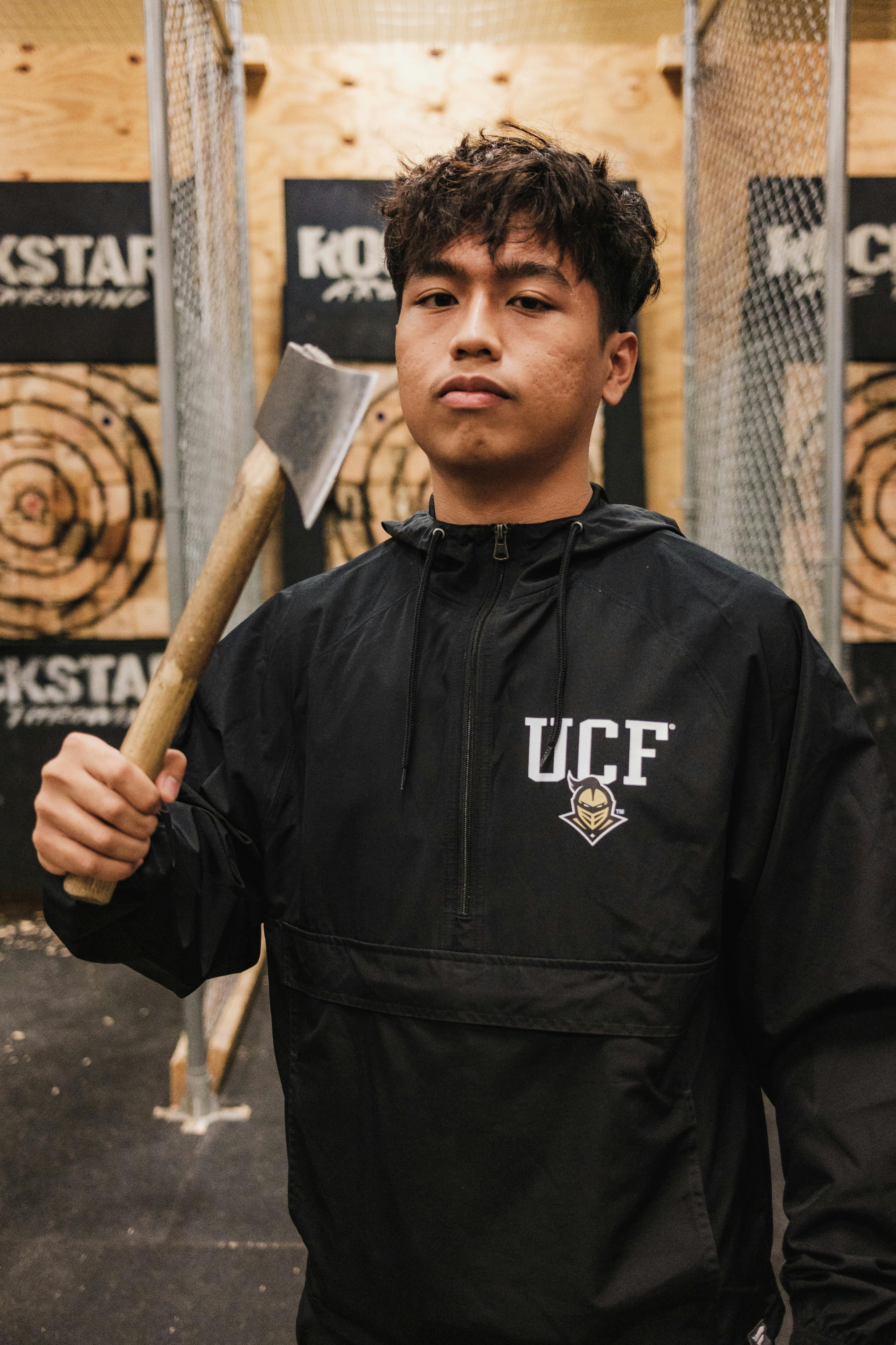 A young person with curly hair smiles at the camera while sitting in front of a laptop against a dark background.