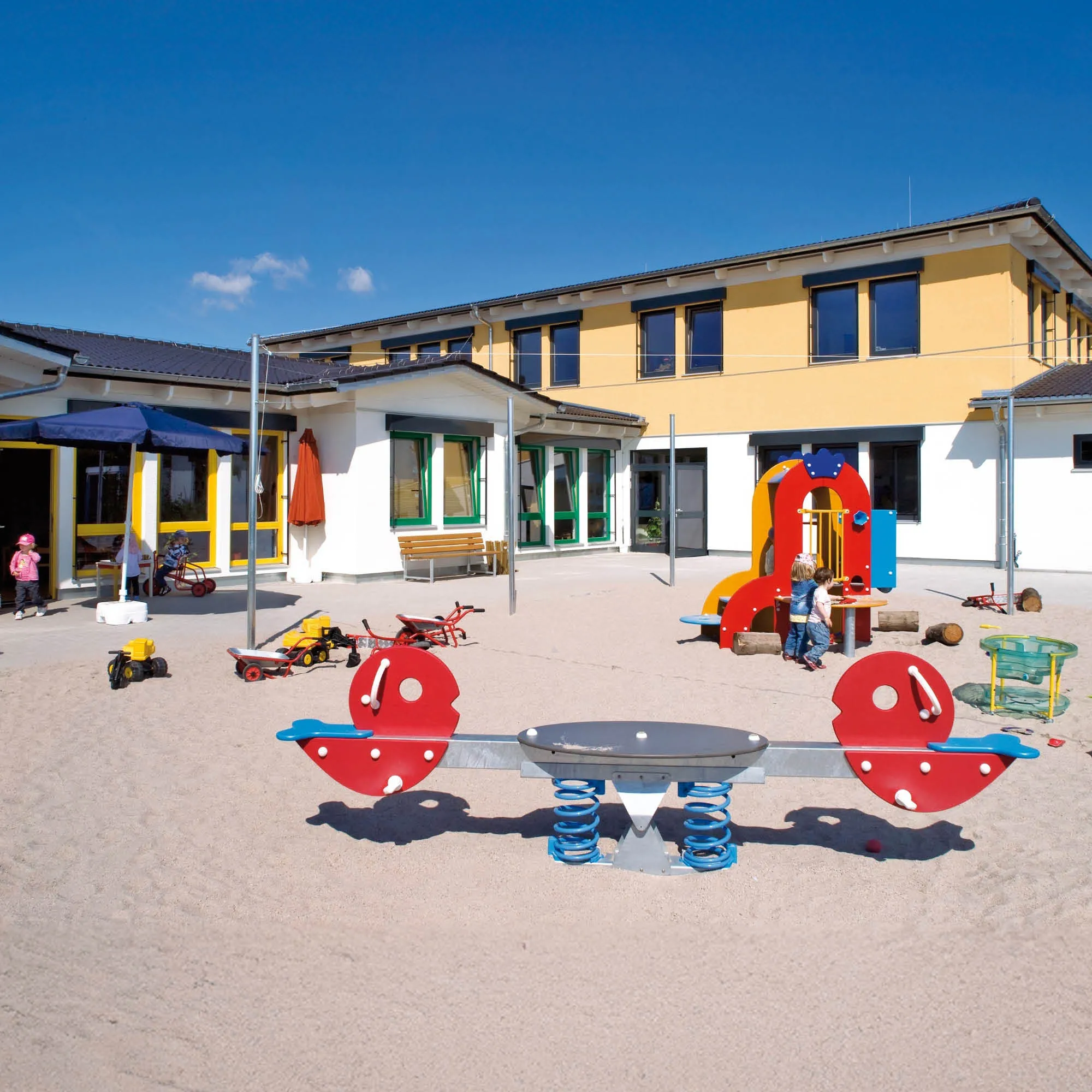 Kindergarten mit einem Sandkasten, Spielgeräten, einer Rutsche und mehreren Kindern im Spiel.