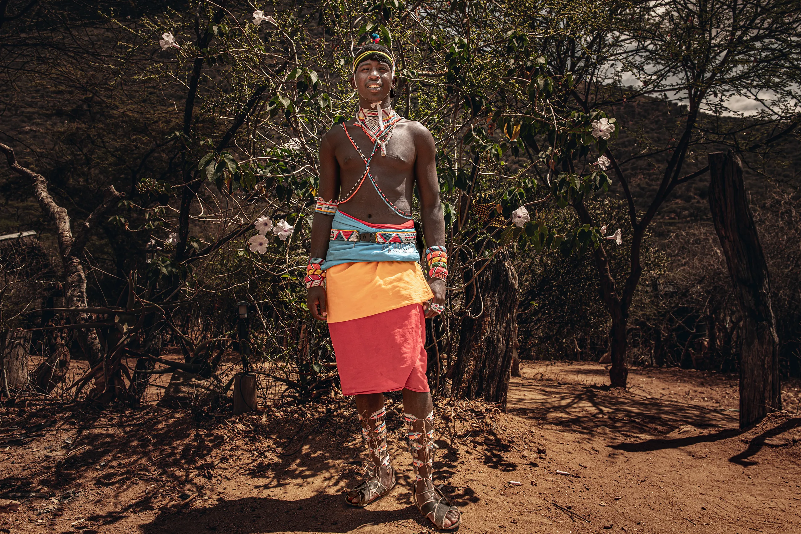 A full body portrait of a Samburu man in traditional dress standing in the dry natural landscape.