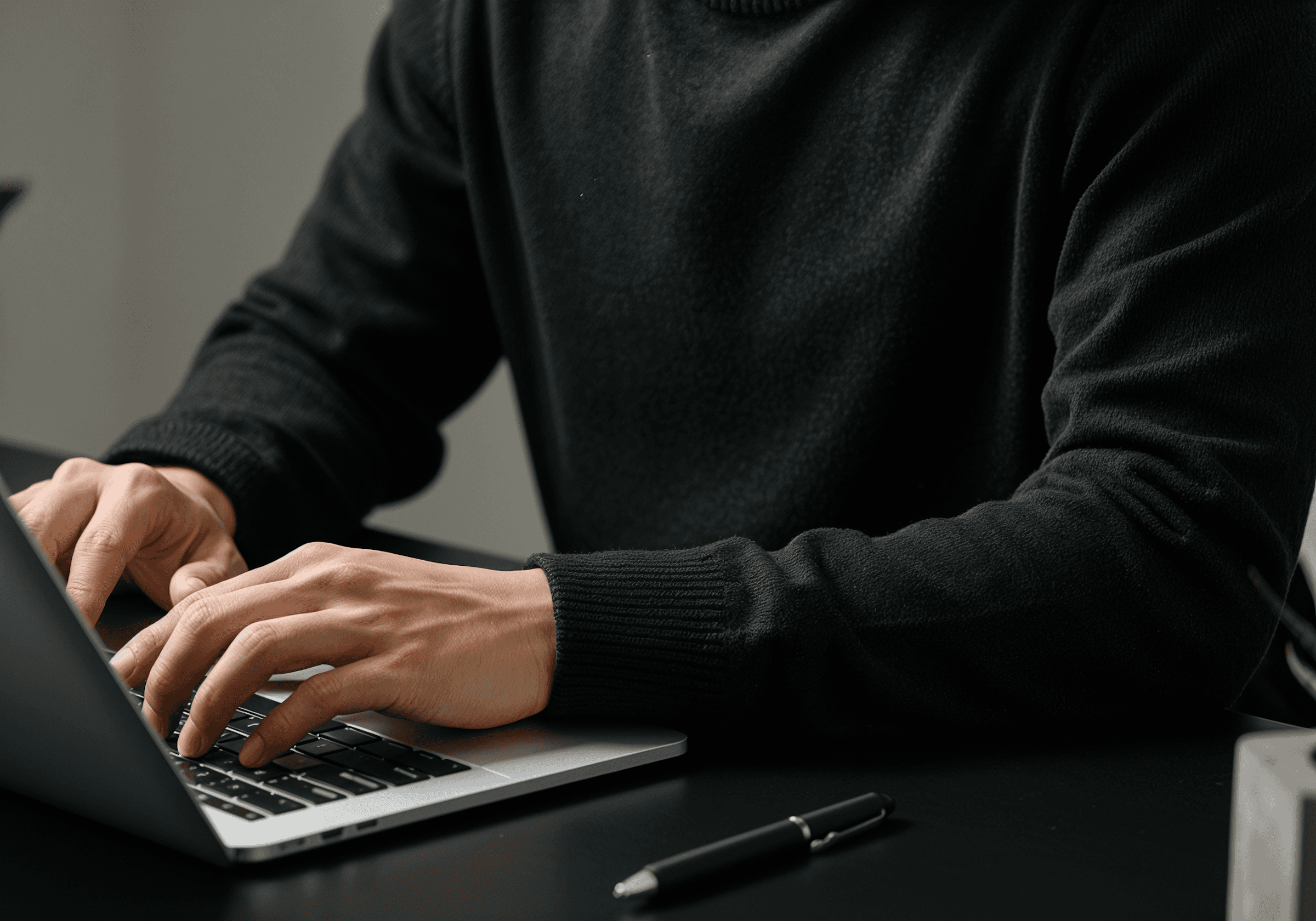 A person in a black sweater typing on a laptop at a black desk, with a pen nearby, creating a professional and focused atmosphere.