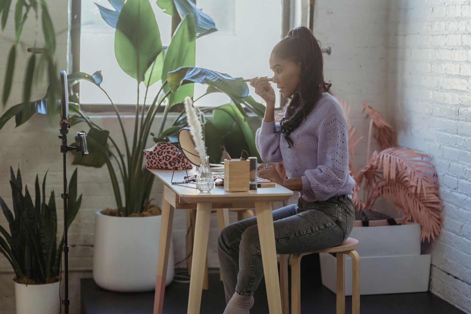 Side view of African American woman with long hair in trendy clothes sitting at table with cosmetics and applying makeup while filming vlog in light room