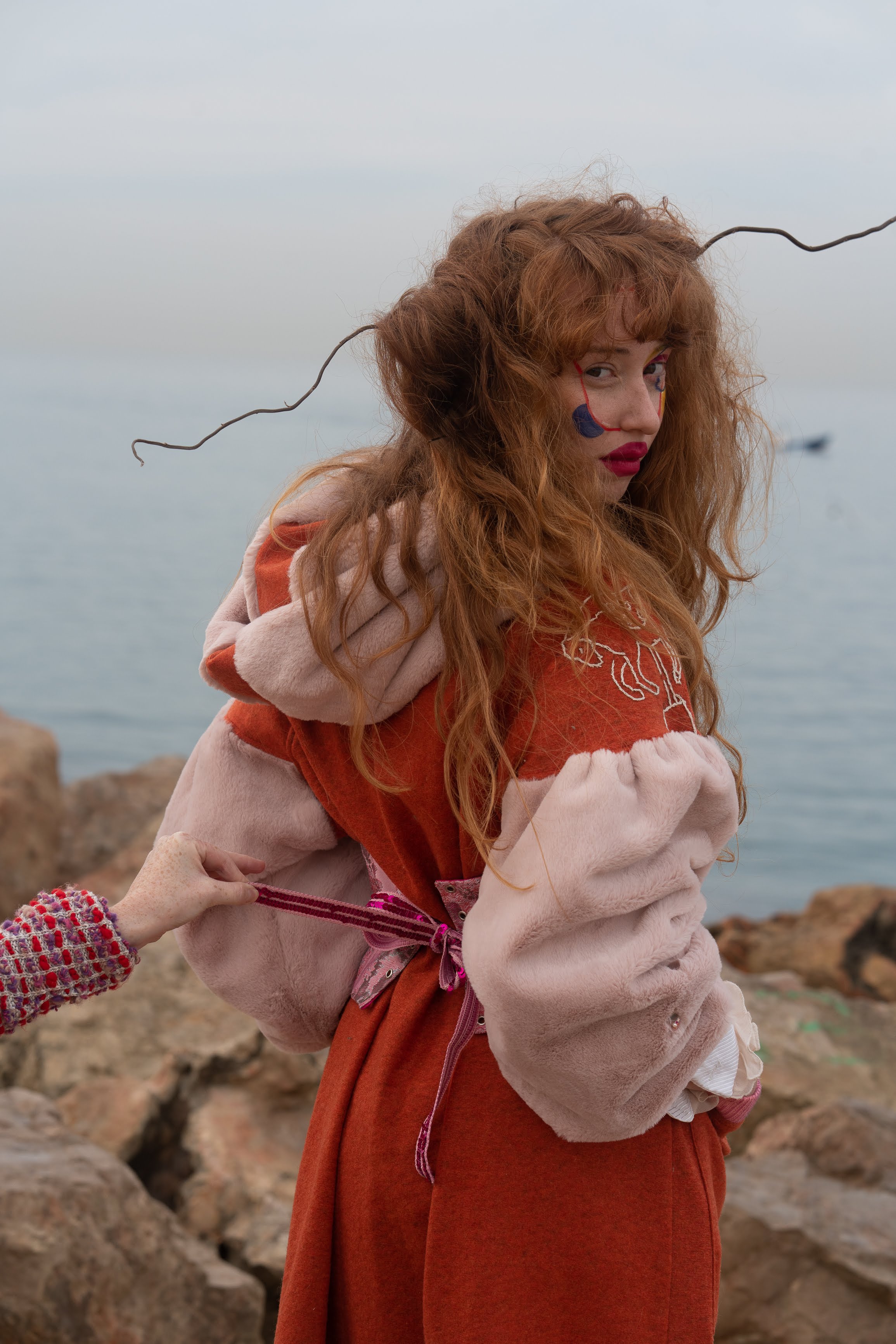 A red haired woman with colorful makeup looking over her shoulder and a hand pulling a ribbon from her dress