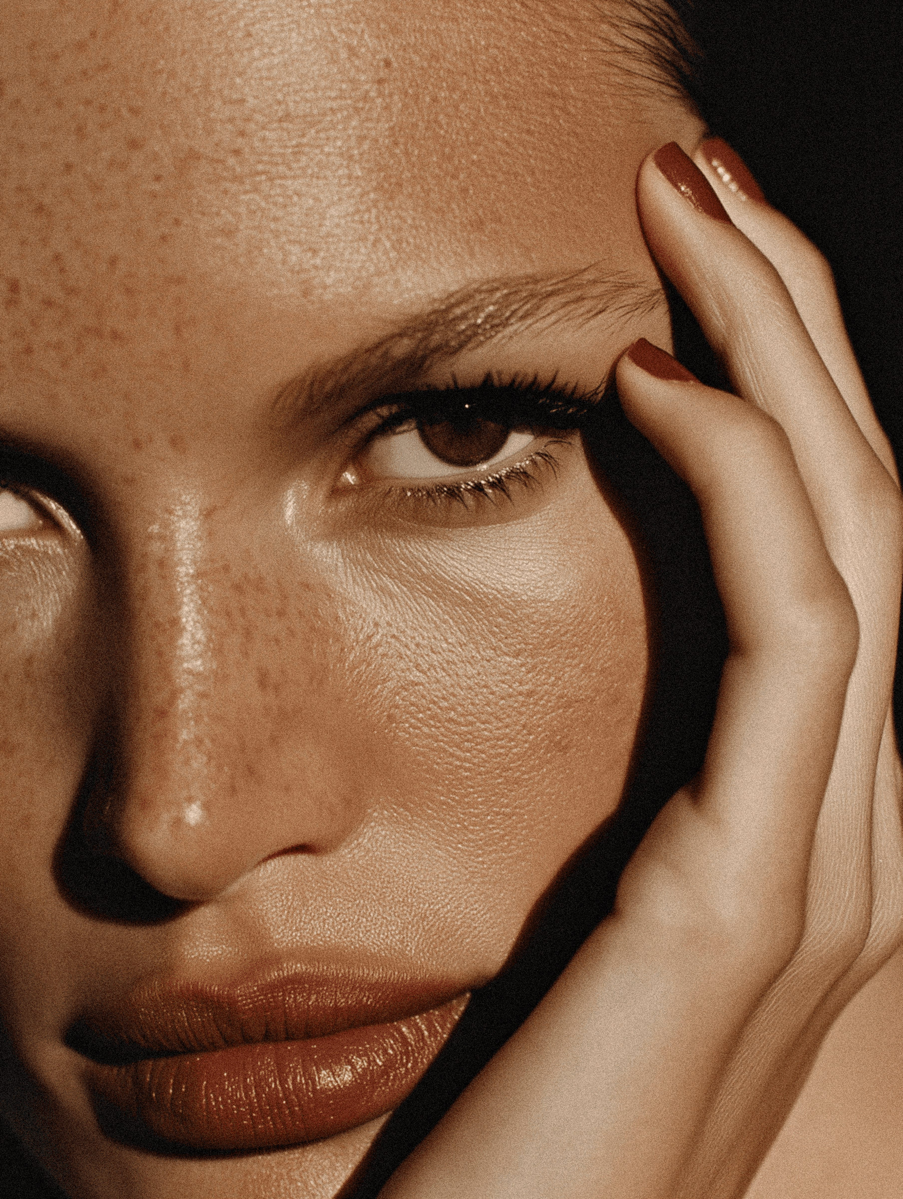 Close-up of a freckled face with brown eyes, matte lips, and red nails, showcasing natural beauty and texture in dramatic lighting.