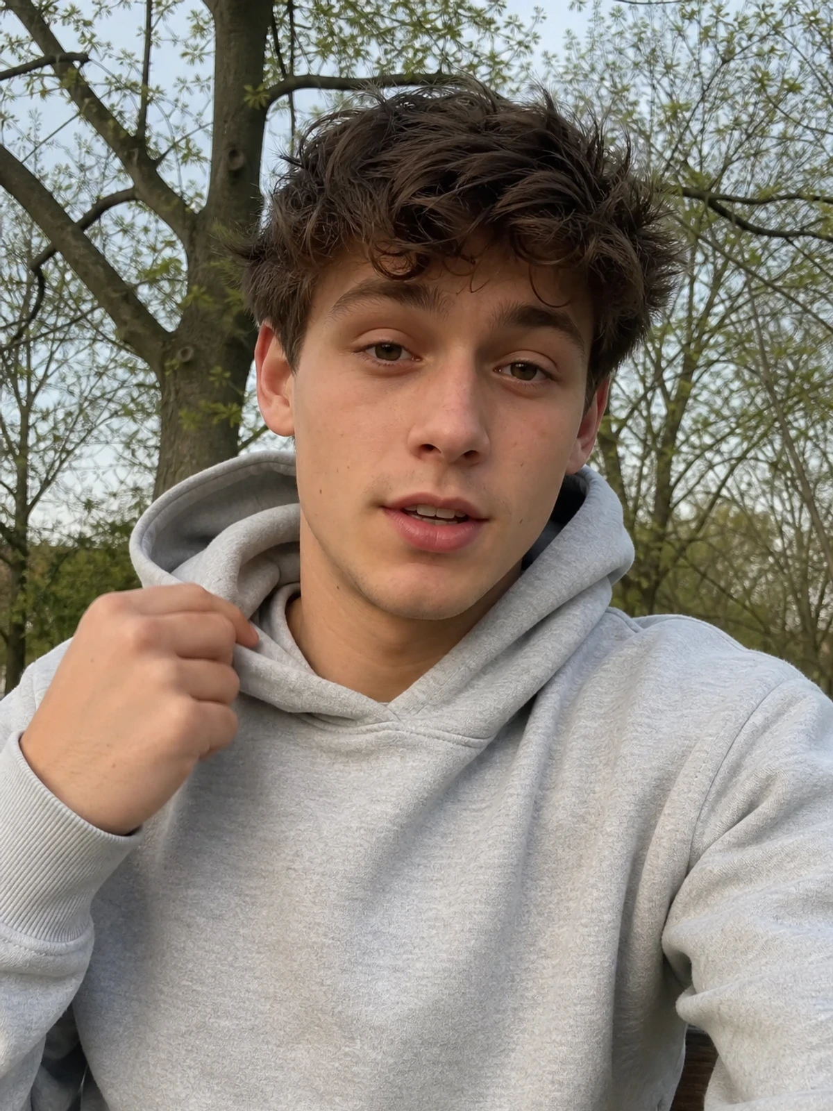 UGC-style photo: young male creator, on a park bench, trying on a plain hoodie while talking to camera, close vertical talking-head shot.