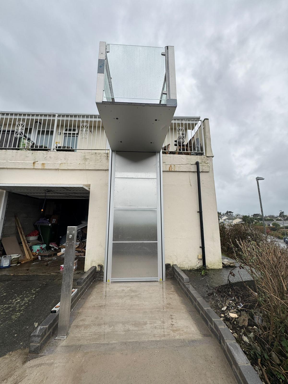 Outdoor vertical platform step lift raised to upper balcony level at a coastal residential property — full stainless steel column and glazed platform enclosure visible against rendered property wall