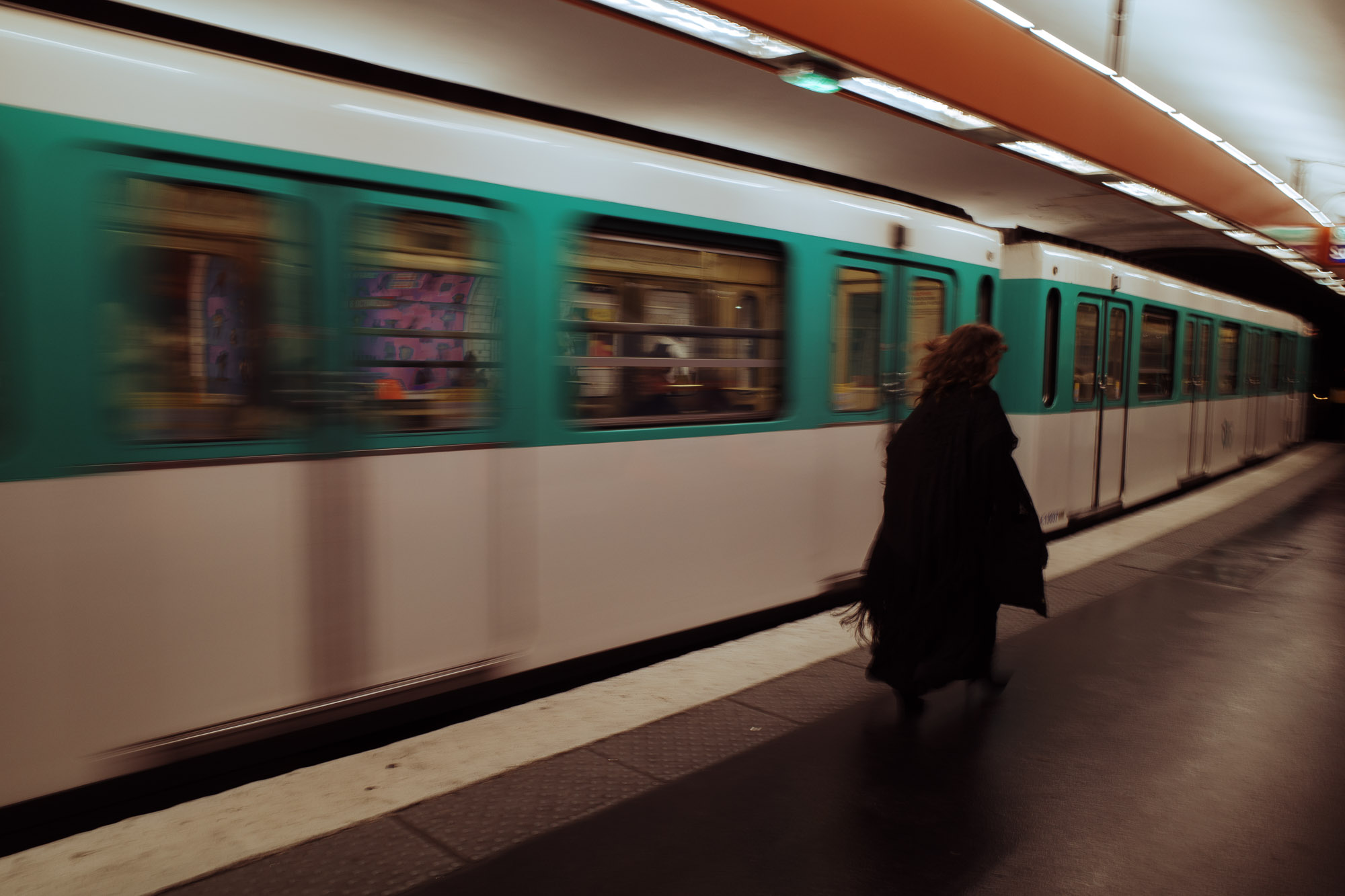 A person in a long coat walks briskly along a platform as a teal and white metro train speeds past in a well-lit underground station, capturing a dynamic urban scene.