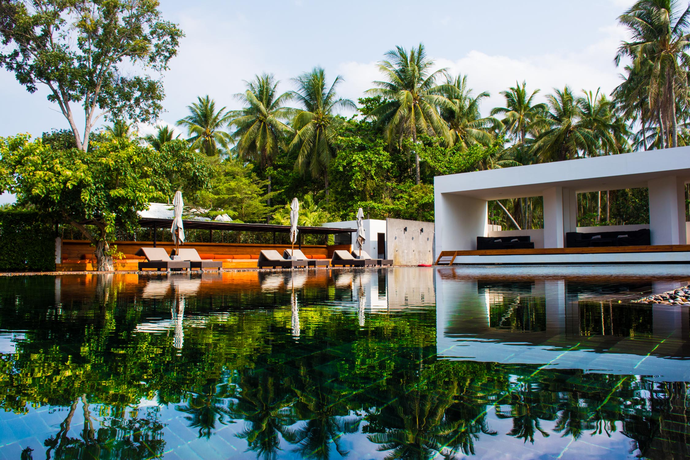 A luxury pool area with white lounge chairs and a modern pavilion reflected in still water under palm trees.