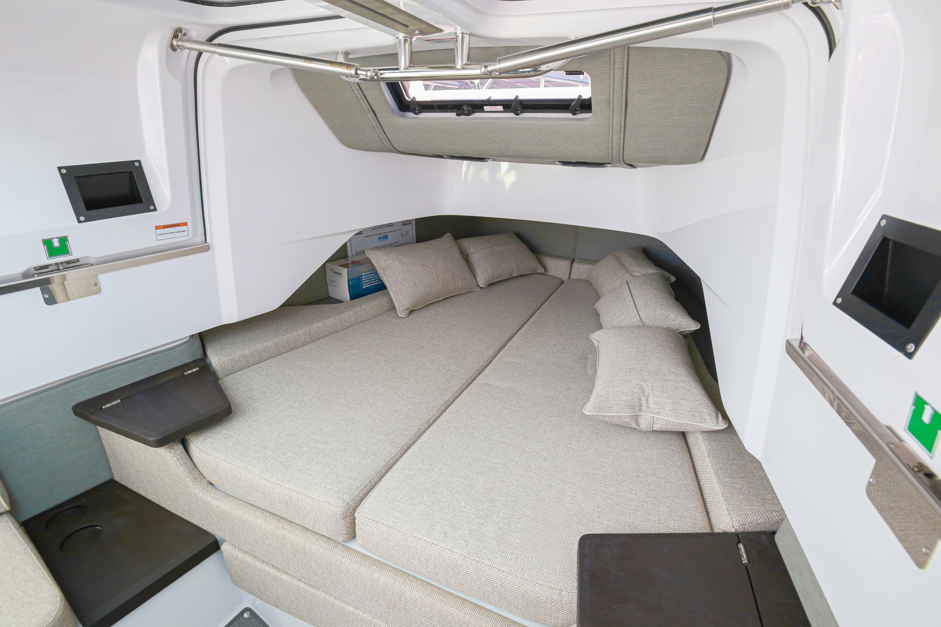Modern yacht cabin interior with beige cushioned seating area, overhead hatch, and built-in entertainment screens on white walls.
