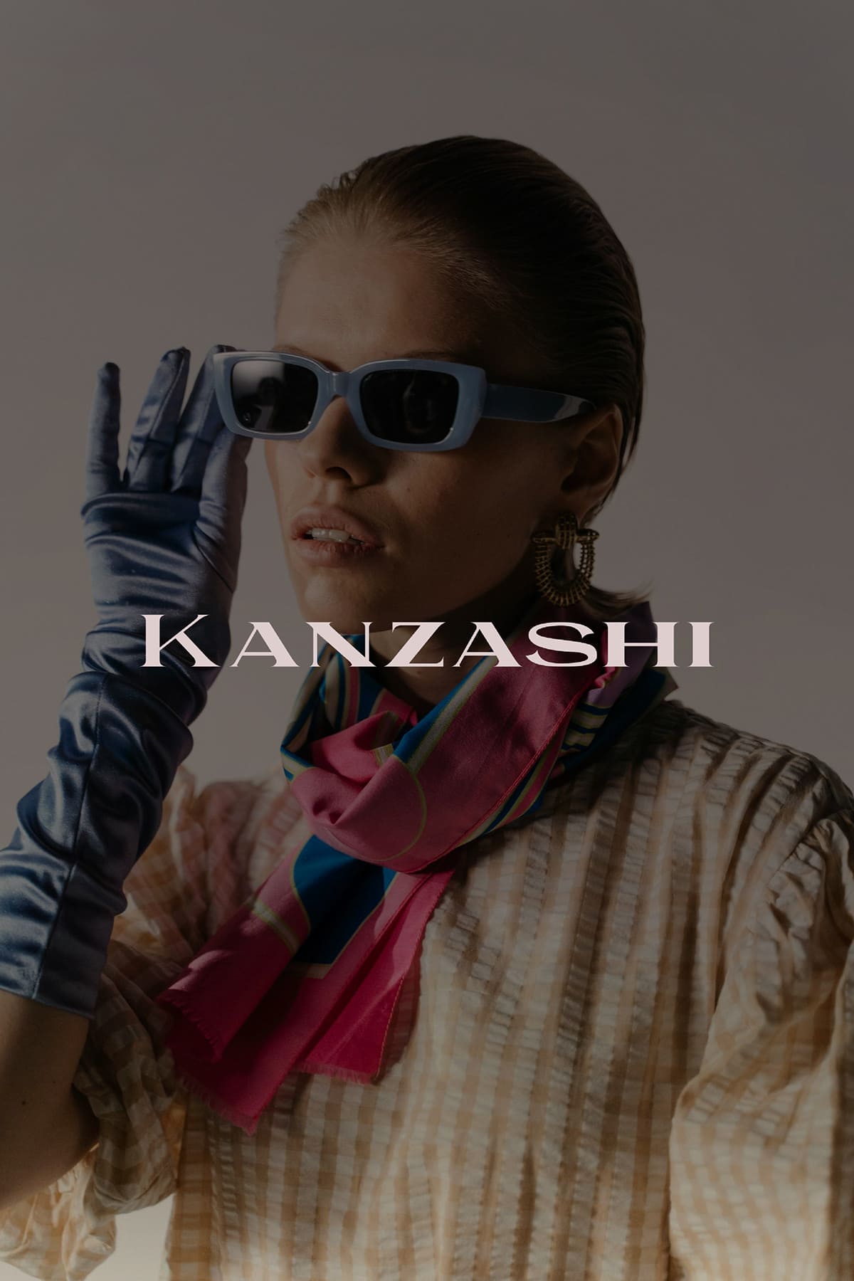 A model with slicked-back hair wears light blue sunglasses and an elegant silk scarf featuring pink and blue patterns. She poses with her left hand raised, adorned in a shiny blue glove. Her outfit consists of a textured striped blouse. The background is softly lit, creating a sophisticated atmosphere, with the word Kanzashi overlaying the image.