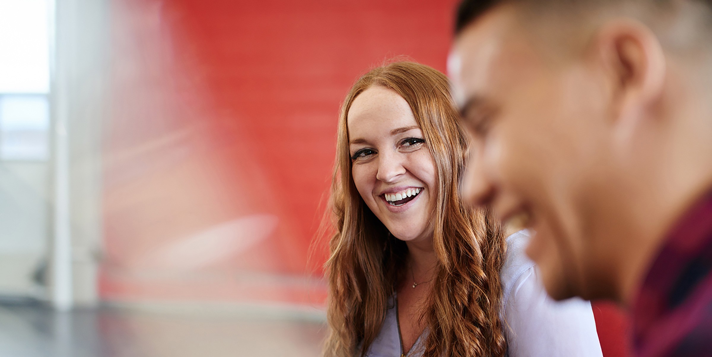 A woman smiling warmly during a conversation with a colleague, with a red wall in the background.