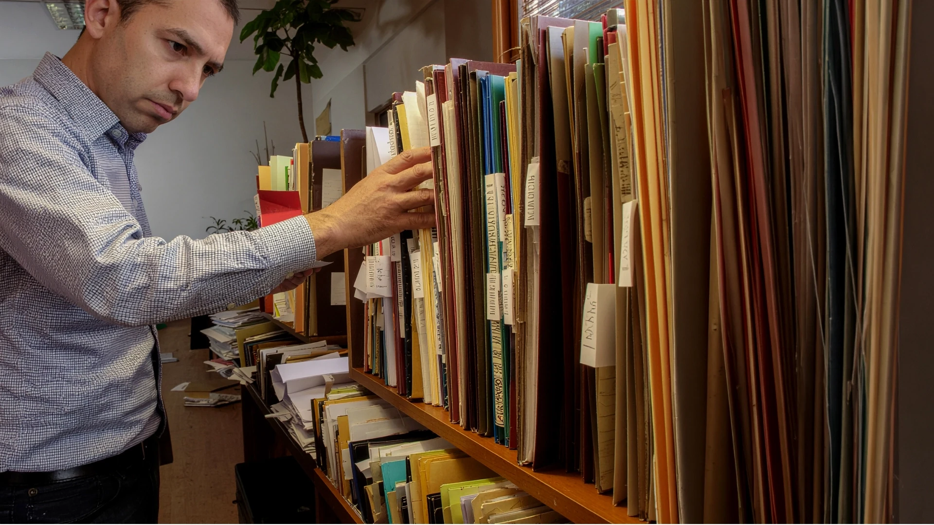 A man searching through densely packed folders and binders on office shelves, looking for a document