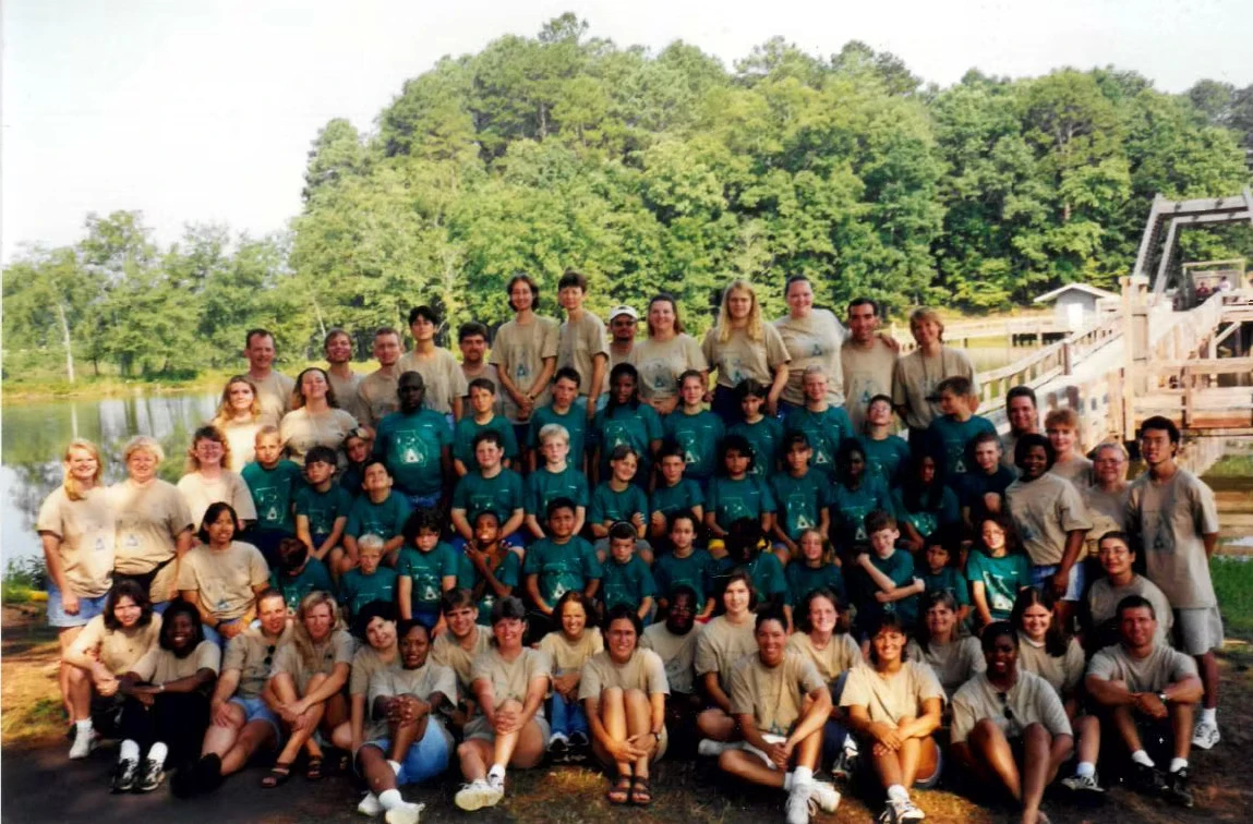 Large group photo of children and adults at Camp Juliena. They are wearing teal and tan t-shirts. A lake and woods are in the background.