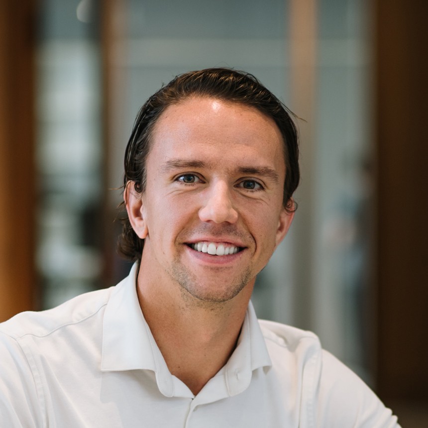 A smiling individual wearing a light shirt, posing for a photo in an indoor setting with blurred background.