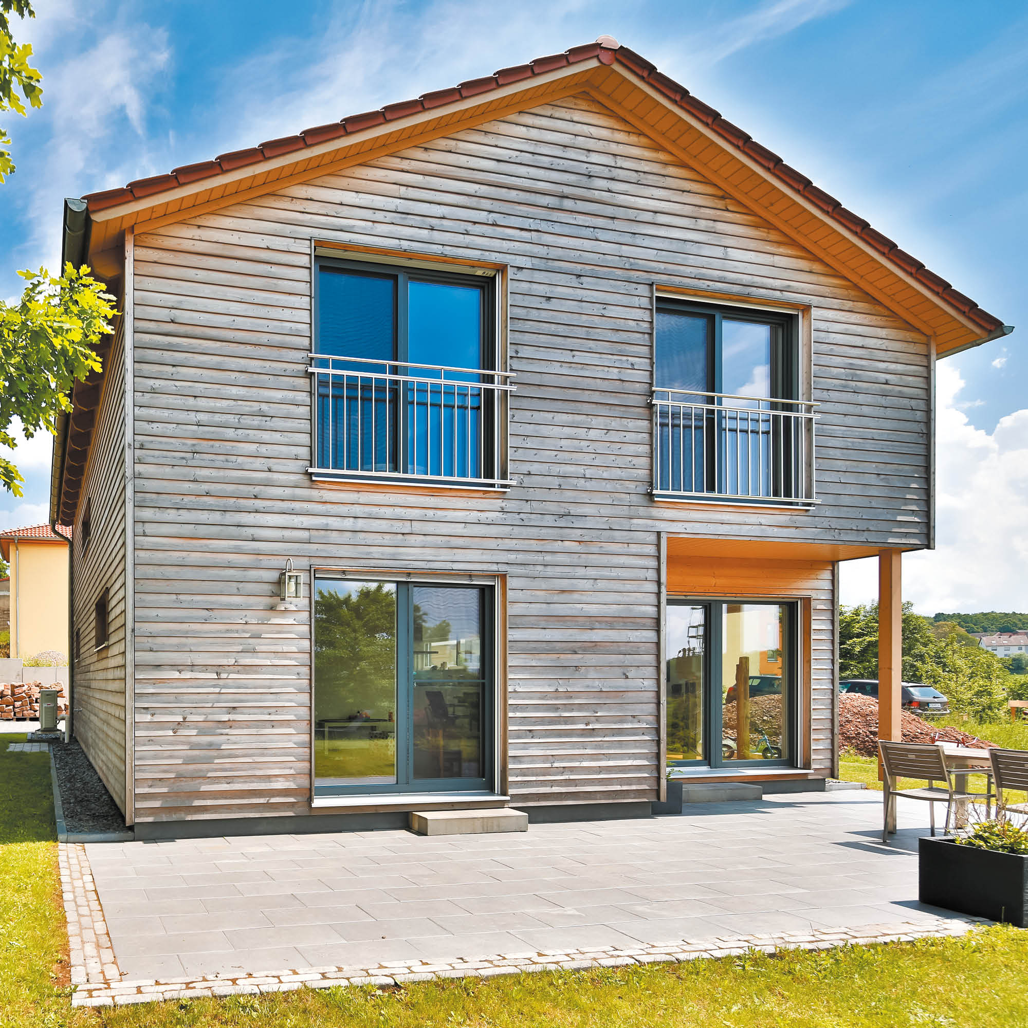 Zweitüriges Holzhaus mit roten Ziegeldach, großzügigen Glasflächen, Balkonen und gepflasterter Terrasse.