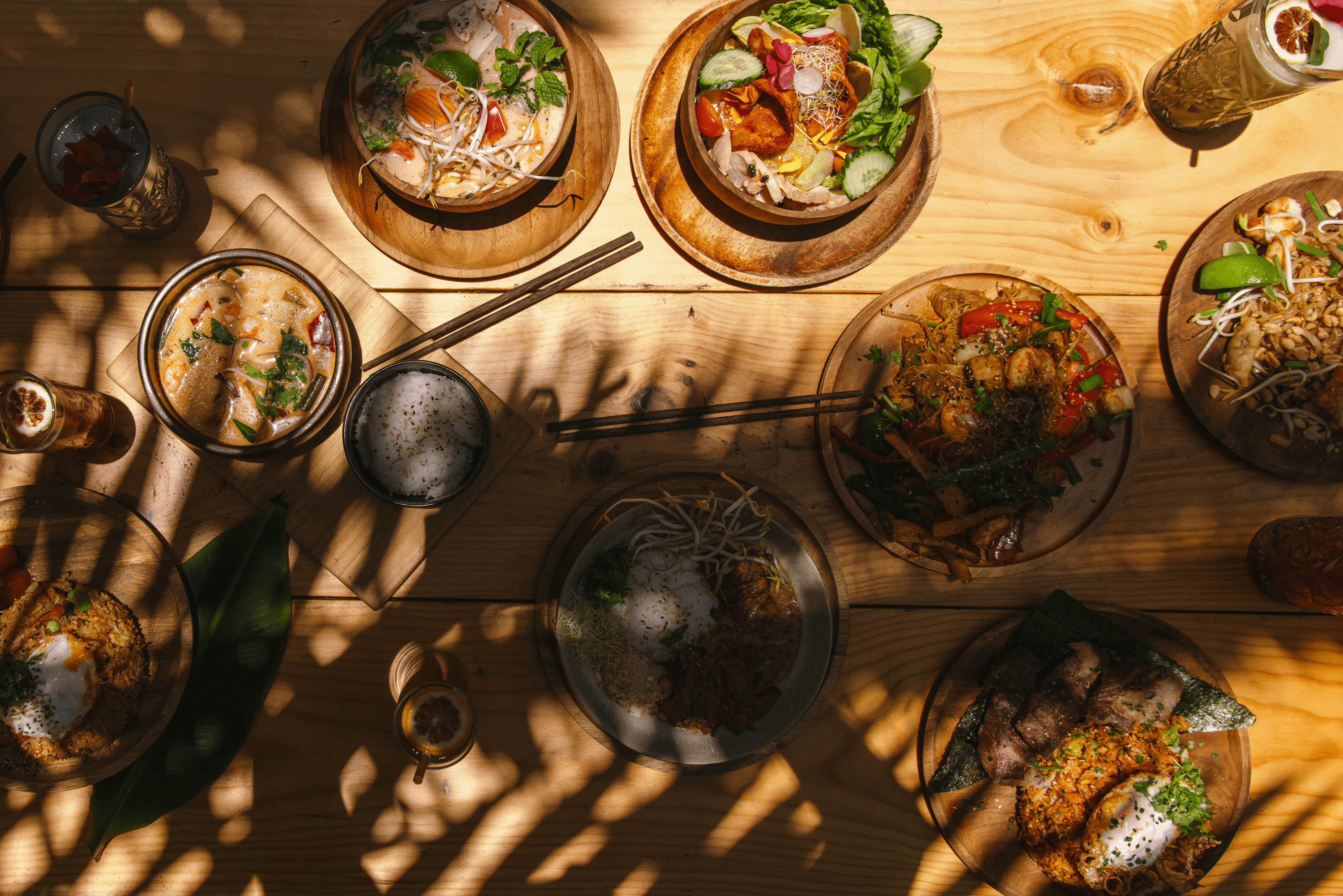 Alt tag: Flat lay of Asian fusion dishes and bowls on a wooden table with palm leaf shadows at Secret Garden, Lajares, Fuerteventura.