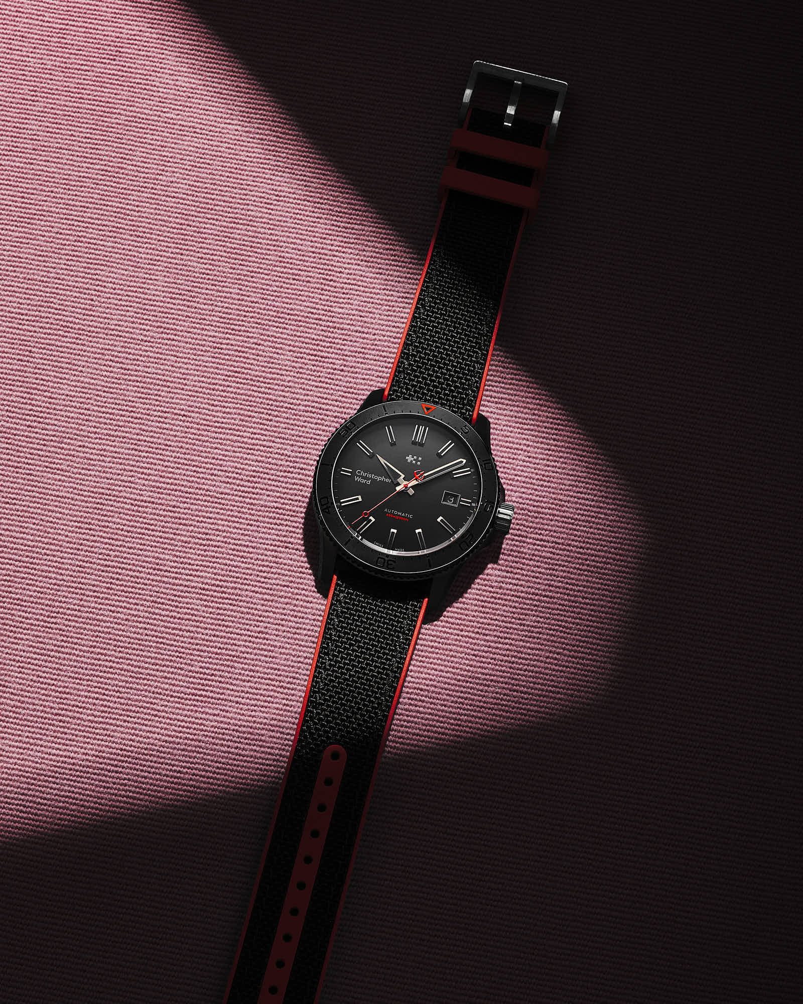 A black wristwatch lies on a pink surface, illuminated by a diagonal beam of light.