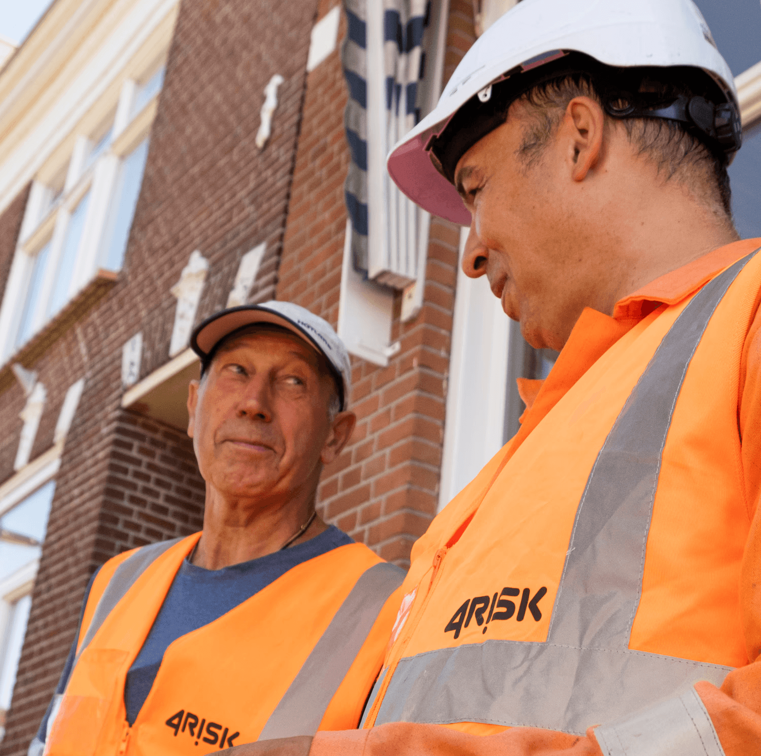 A smiling couple takes a selfie under a clear blue sky. The woman wears a red jacket, and the man, in a green top, holds a smartphone.