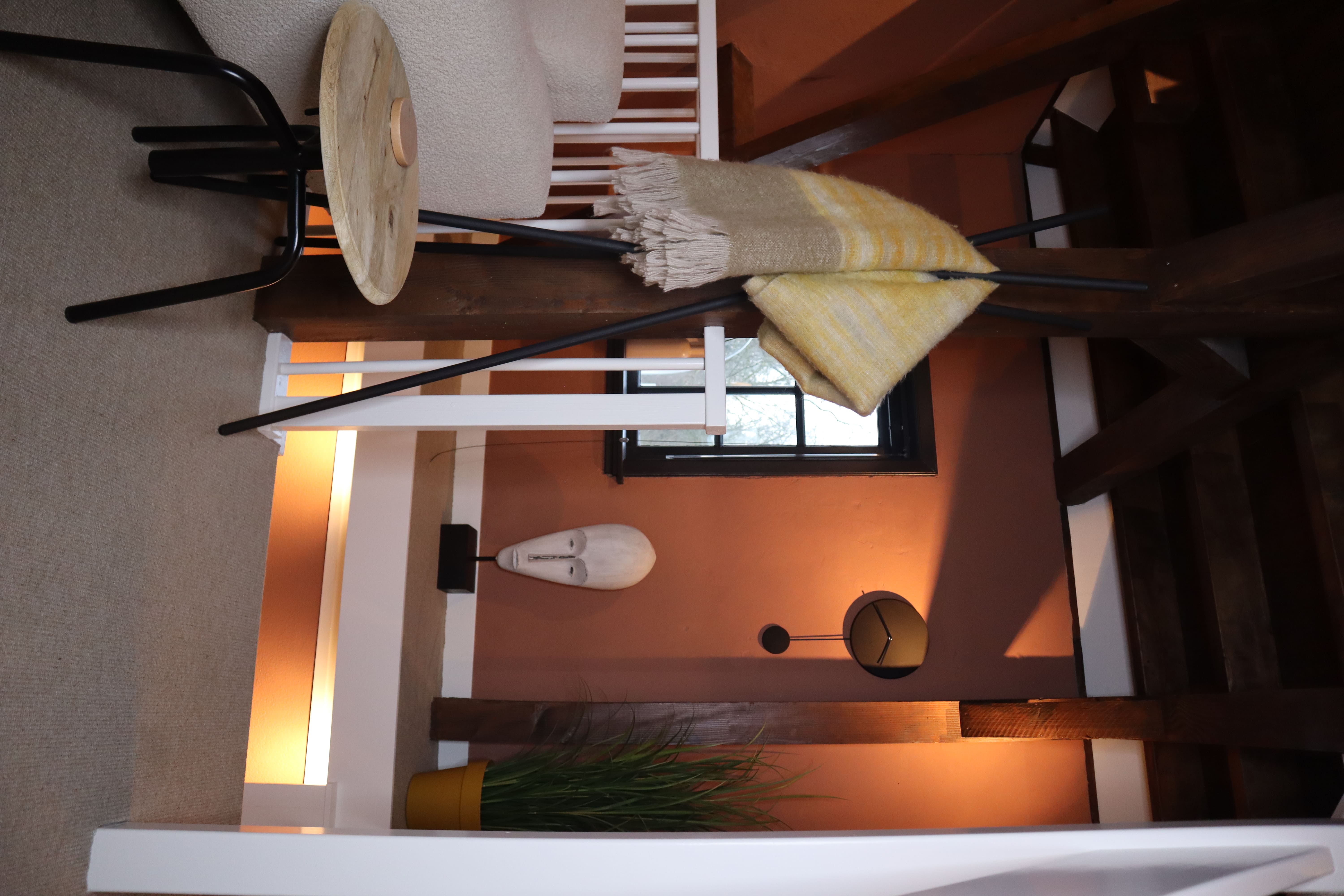 Hoek van een woonkamer met een decoratieve witte sculptuur, een plant in een gele pot, een gebreide deken aan een houten paal en een kleine salontafel.