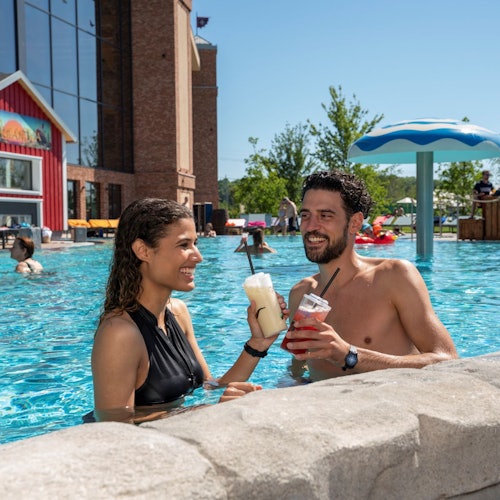 两人在游泳池中微笑,手中拿着饮品。附近还有其他人在游泳。背景是红色建筑和伞形结构。