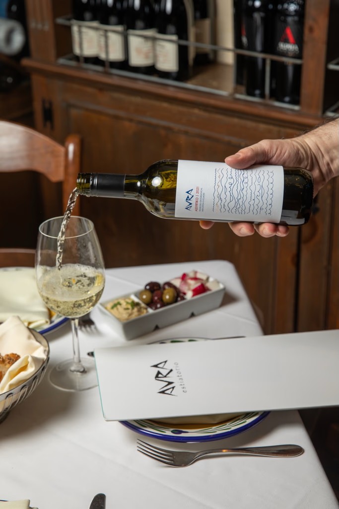 A hand pours wine from a bottle into a glass on a dining table set with plates and cutlery.