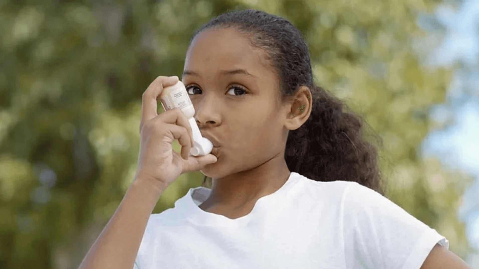 A young child takes a breath from an asthma inhaler with blurred trees in the background