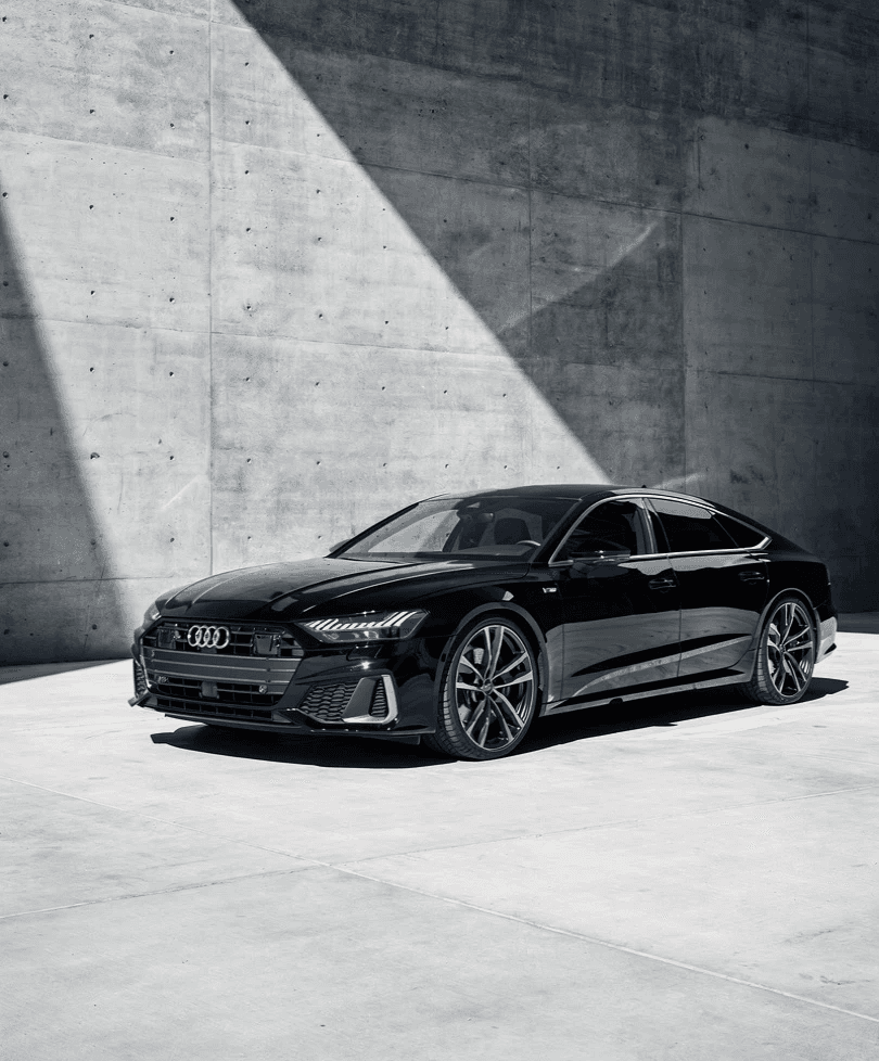Sleek black car in a modern concrete space, partially lit by a diagonal beam of sunlight, creating a dramatic contrast. The scene feels sophisticated and elegant.