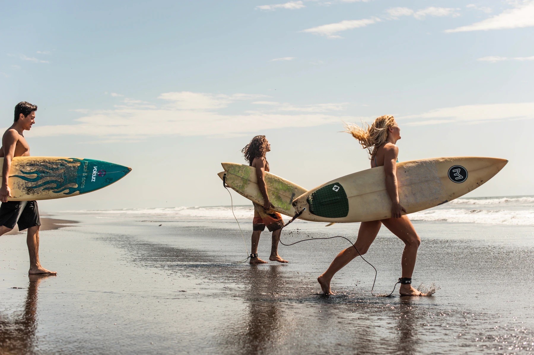 Tres surfistas llevan sus tablas a lo largo de la orilla de una amplia y soleada playa.