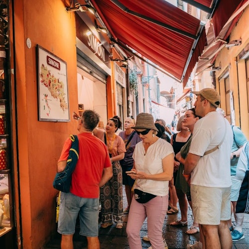 אנשים בבגדי קיץ קלילים מתאספים מחוץ לחנות בסמטה צרה עם קירות כתומים וסוככים אדומים.