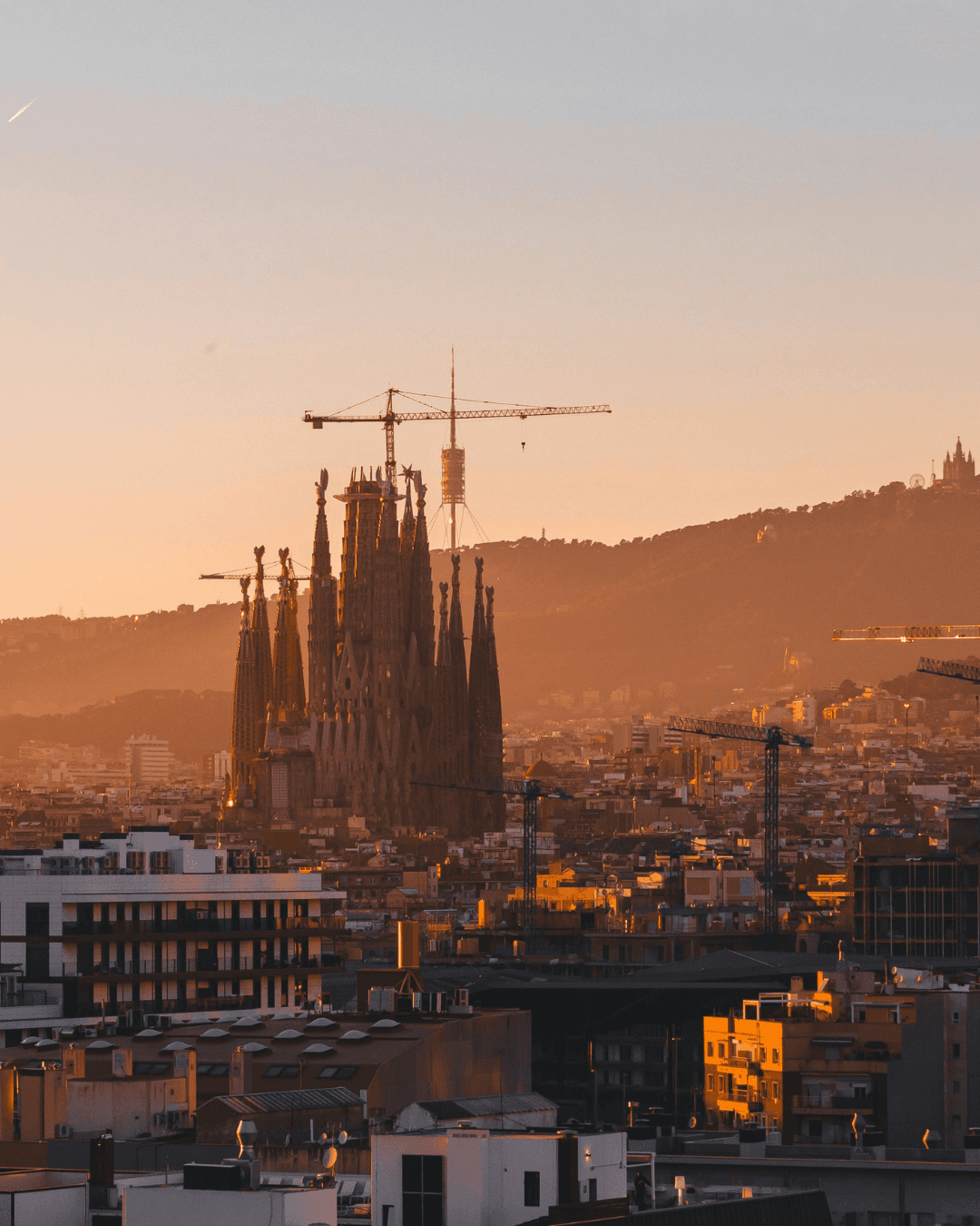 Sagrada Família exterior at sunset with construction cranes visible against the Barcelona skyline.