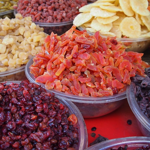 Assorted dried fruit pieces in plastic bowls including slices of papaya, banana chips, yellow chunks, red berries, and black berries.