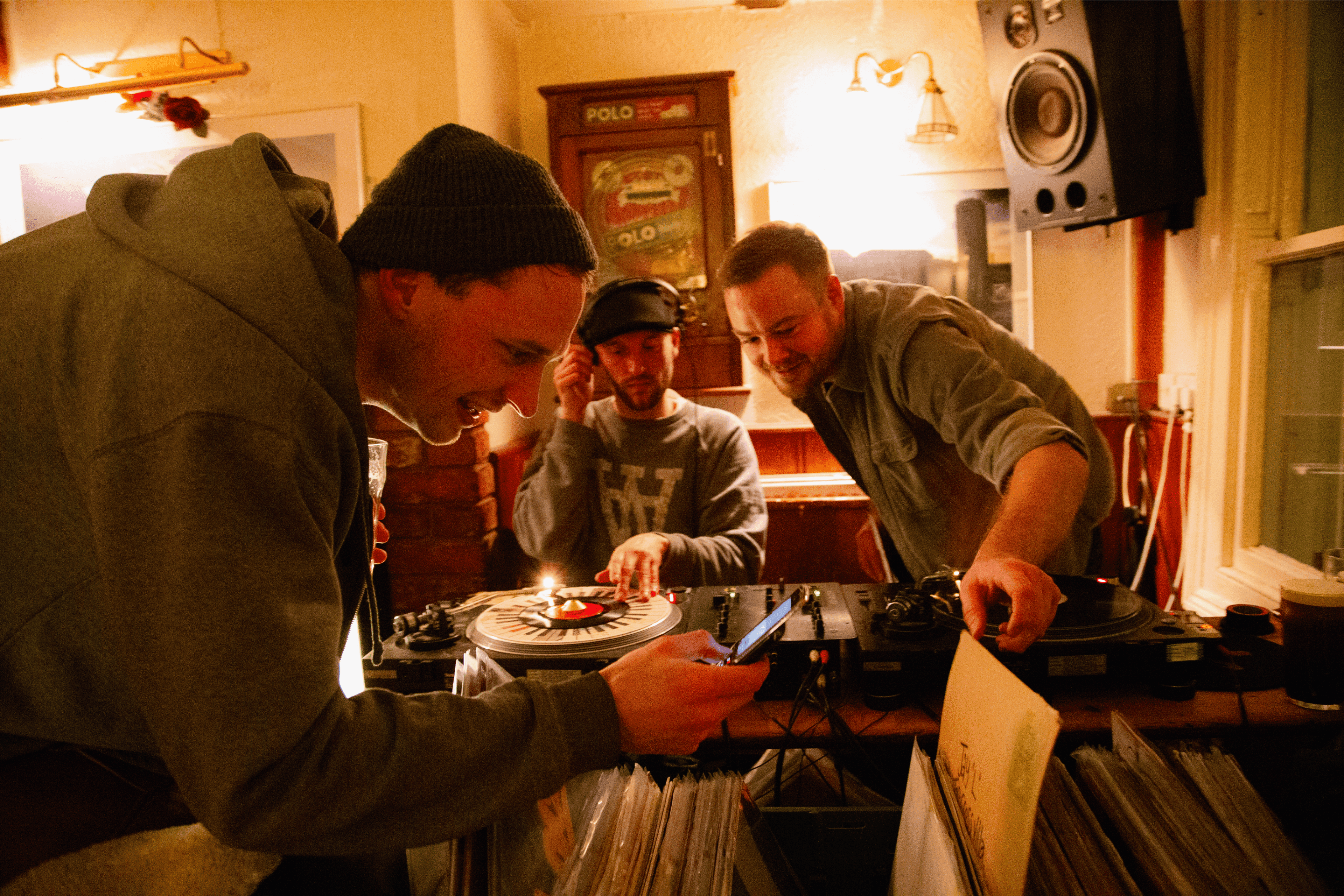 Three figures surround the turntables and bags of records at The Bell pub during a Boundary Object event