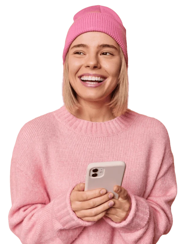 A woman in a pink sweater and beanie smiles while holding a cell phone in her hand.