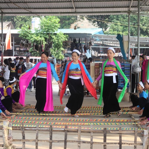 Tre kvinder i farverig traditionel påklædning optræder med en folkedans under et metaltag, omgivet af tilskuere og markedsboder.