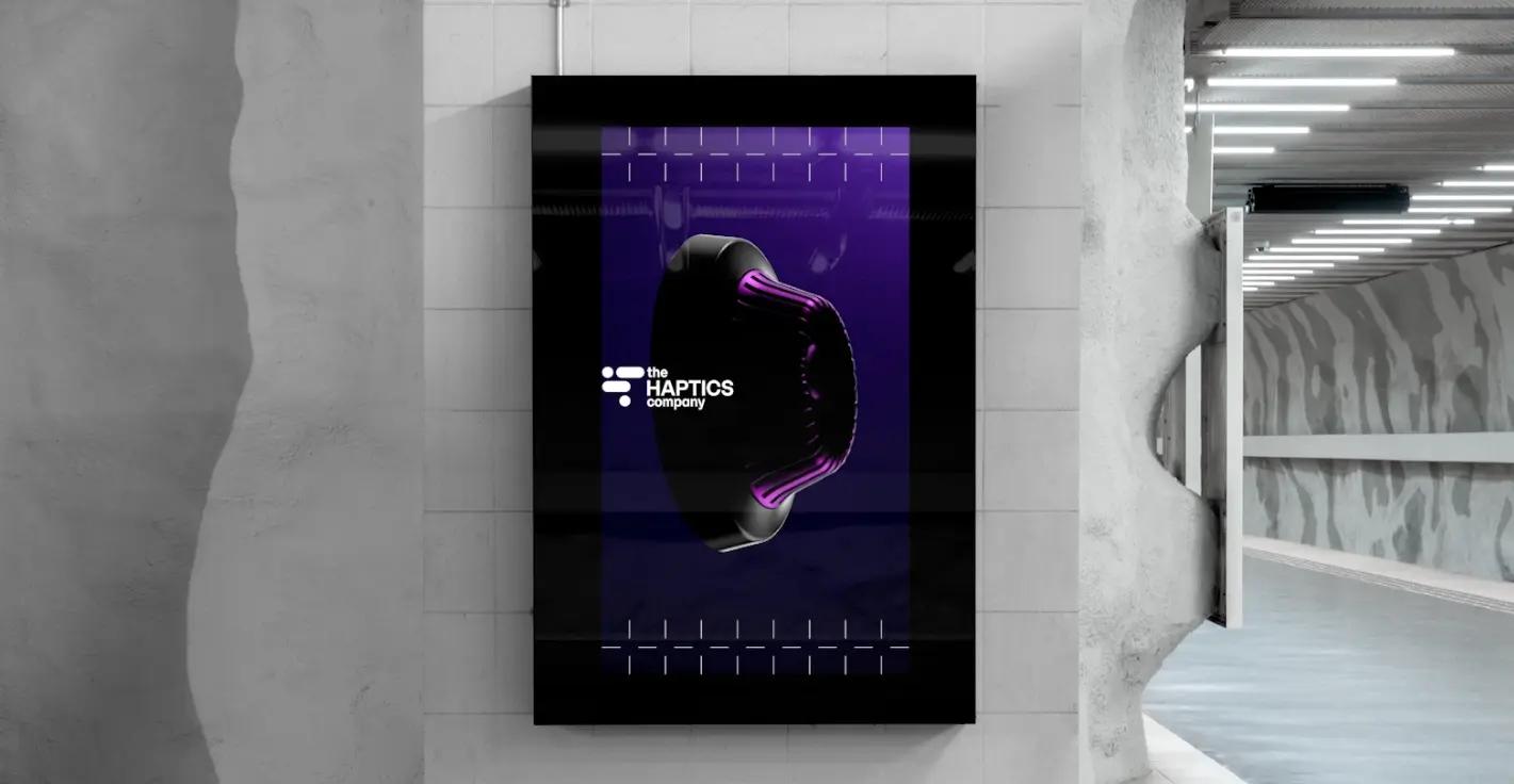Backlit digital billboard in an industrial corridor shows "the HAPTICS company" logo and a purple haptics device, reflecting visual identity tech.