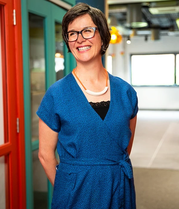 Photo of Marianne O'Loughlin in a blue dress standing in a hallway smiling and looking off to the side of shot