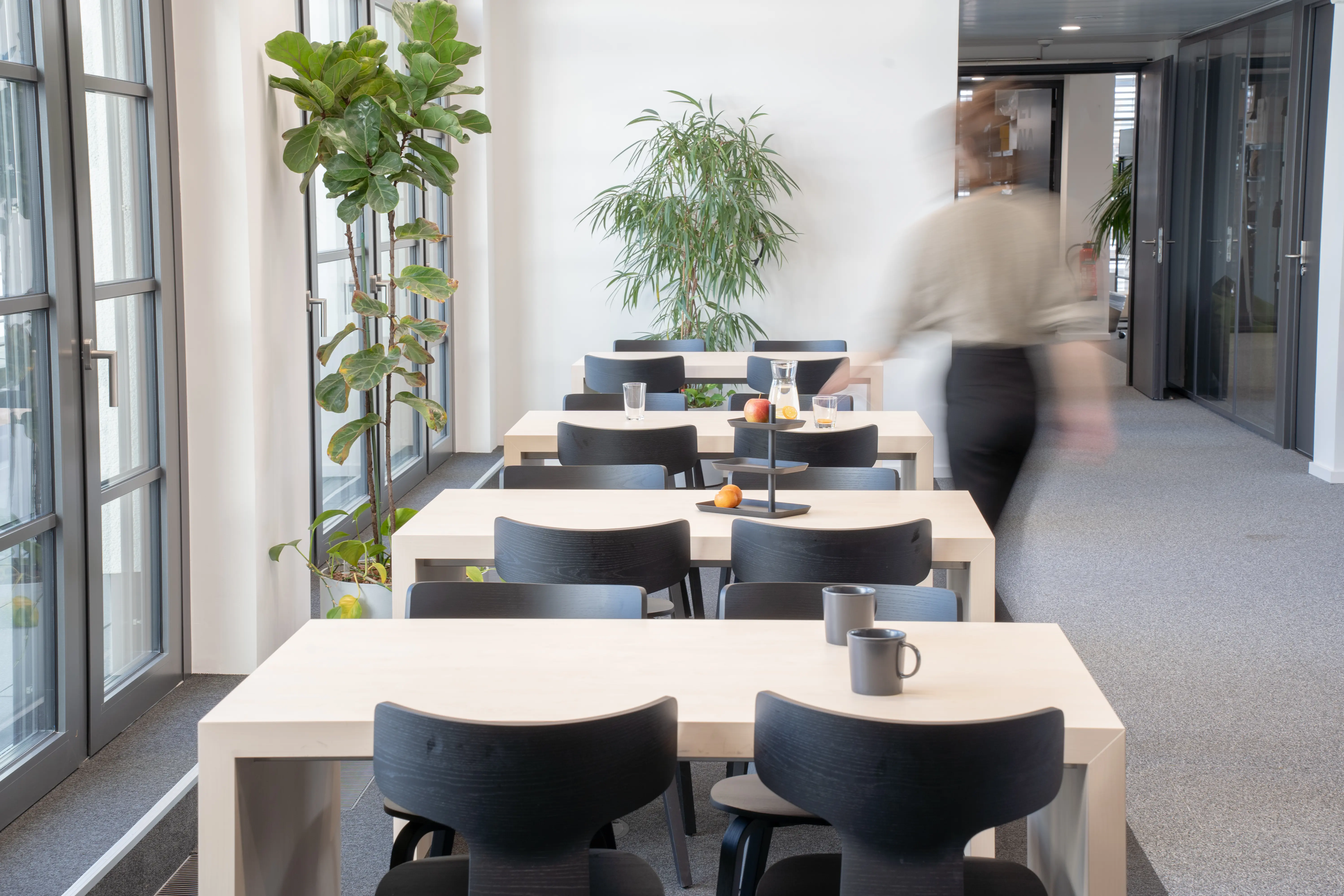 Canteen at AI START-UP MUNICH with pale wooden block tables, black wooden chairs, fruit and ceramic mugs on the tables, large plants and a person walking past in motion blur.