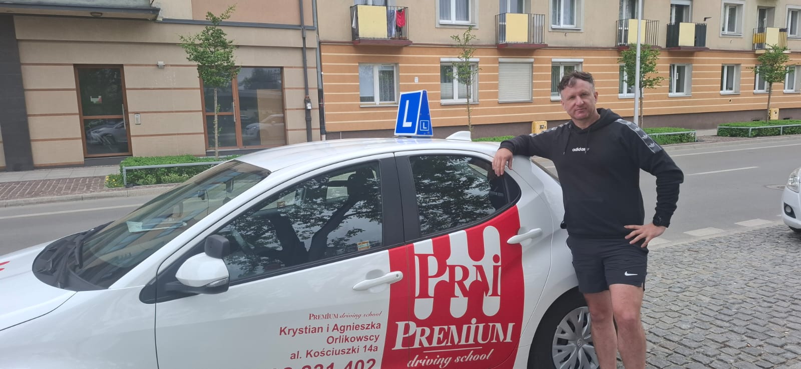 Adam Ślężak – Instruktor z ręką opartą o czerwono-białe auto Premium, w sportowej bluzie.