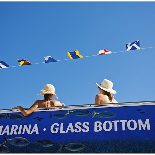 Two people in sun hats on a blue boat with "MARINA GLASS BOTTOM" written on it, under colorful signal flags.