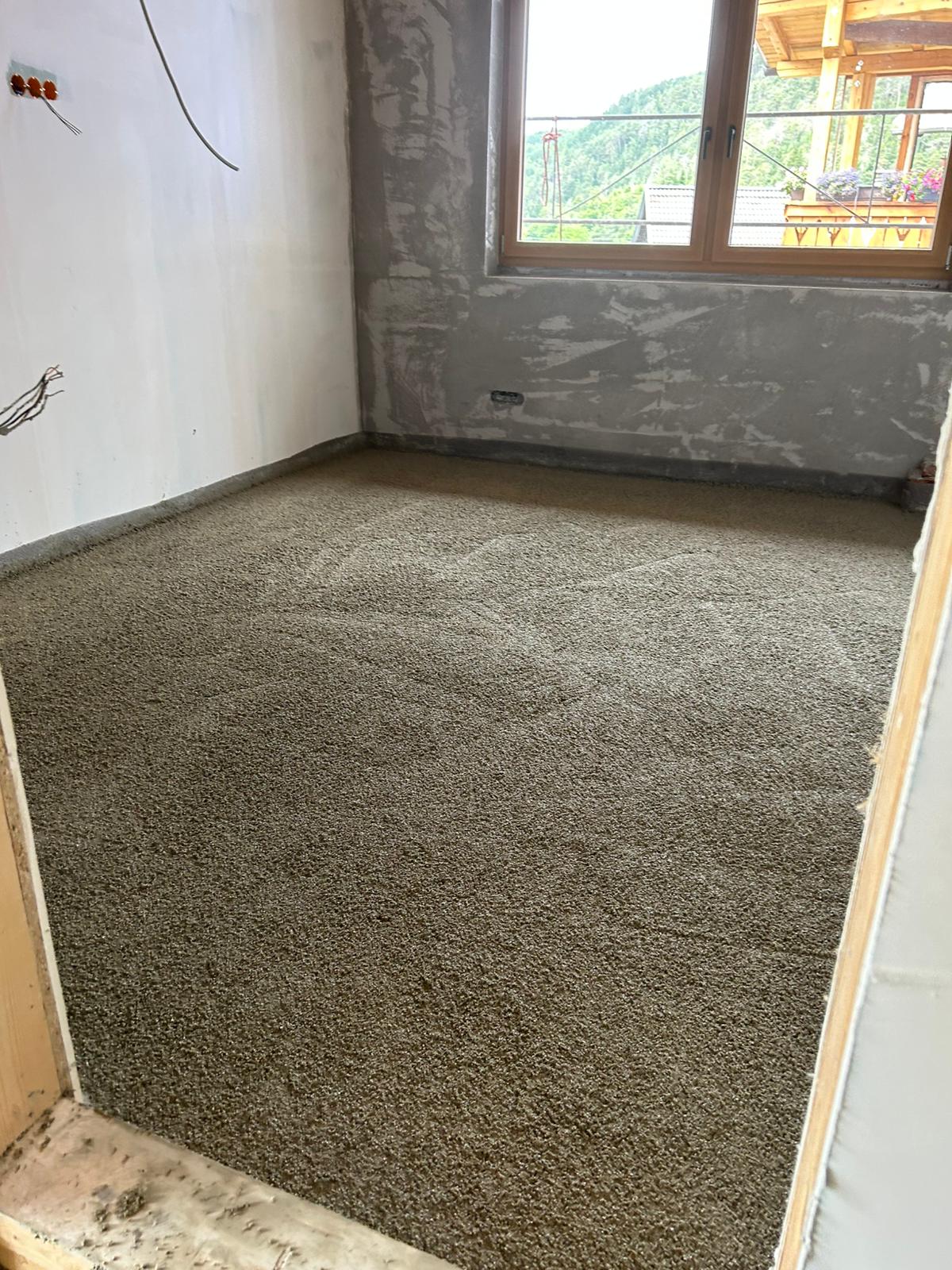 Raum mit frisch gegossenem Betonboden, bereit für den Innenausbau. Im Hintergrund bietet ein Fenster einen herrlichen Bergblick.
