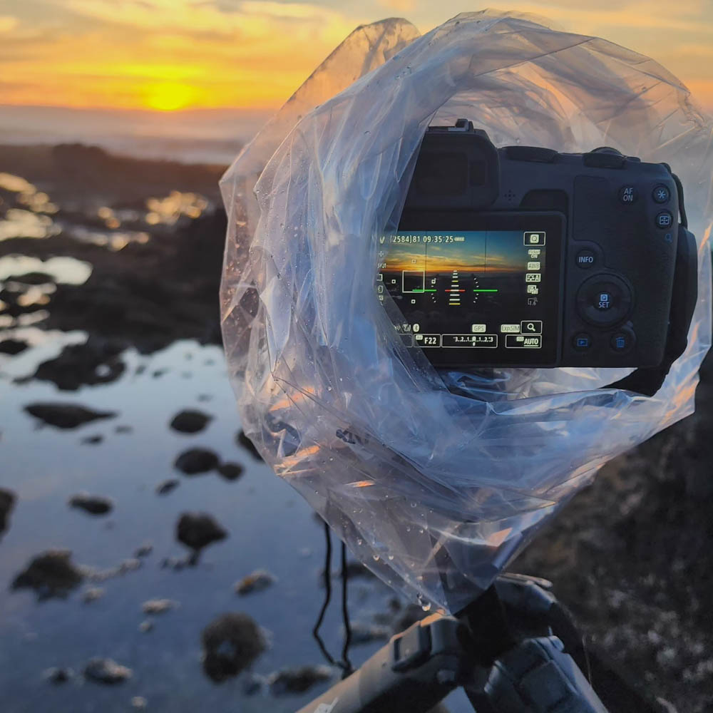 Camera wrapped in plastic on a tripod, capturing a coastal sunrise over tidepools.