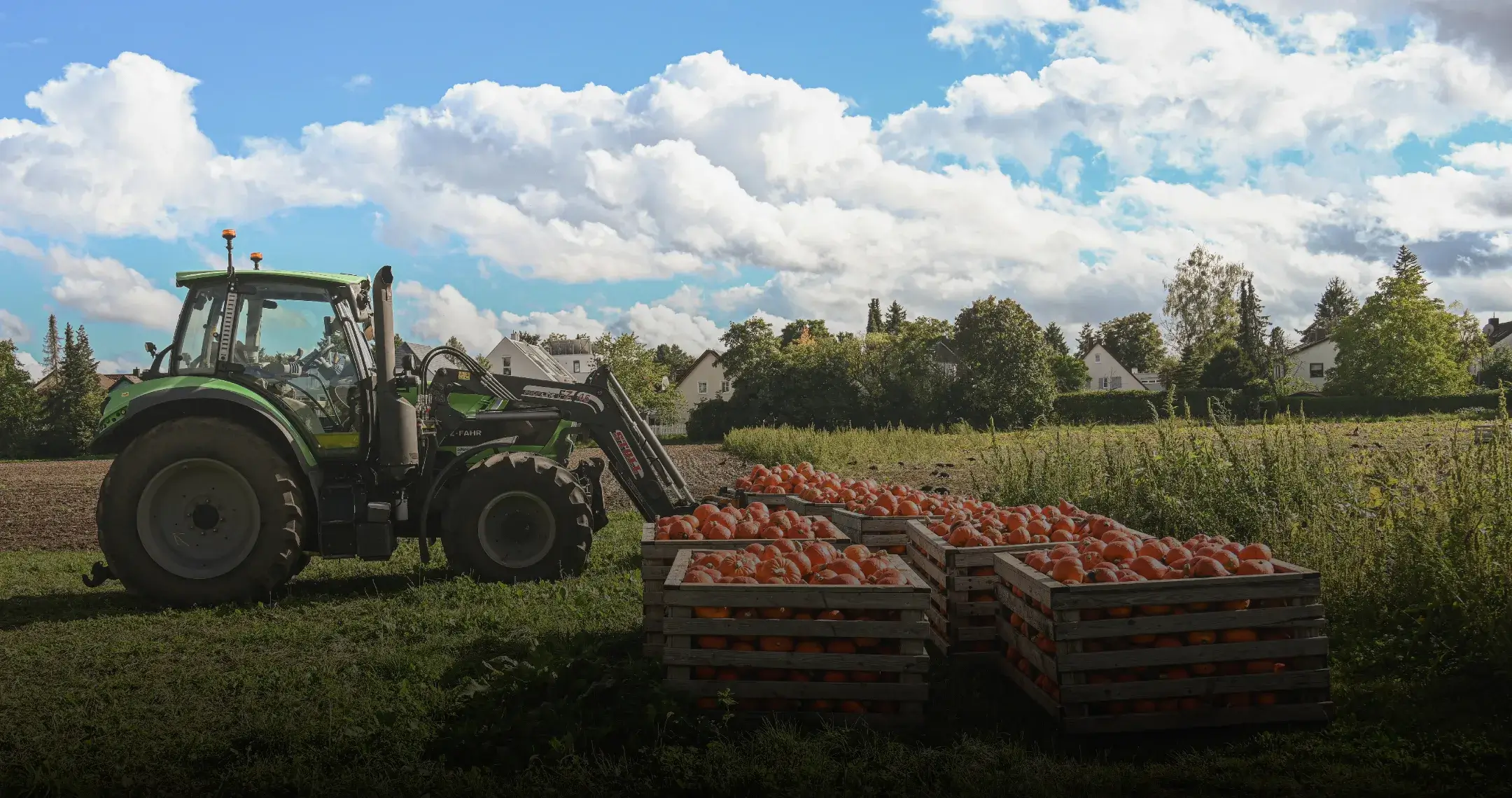 A green tractor is parked in a vibrant field under a partly cloudy sky, next to crates filled with orange pumpkins, evoking a harvest season atmosphere.