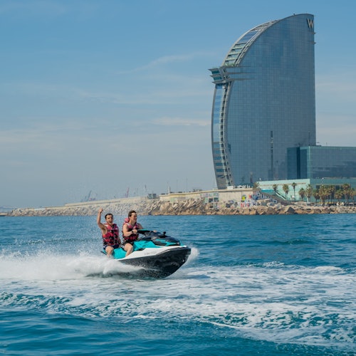 兩個人正在海上騎著水上摩托艇，背景是一棟高聳、弧形的酒店大樓和海岸線。