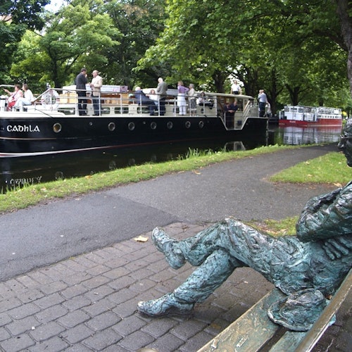 A statue sits on a bench near a canal where a boat with passengers is moored. Trees and a paved path are in the background.