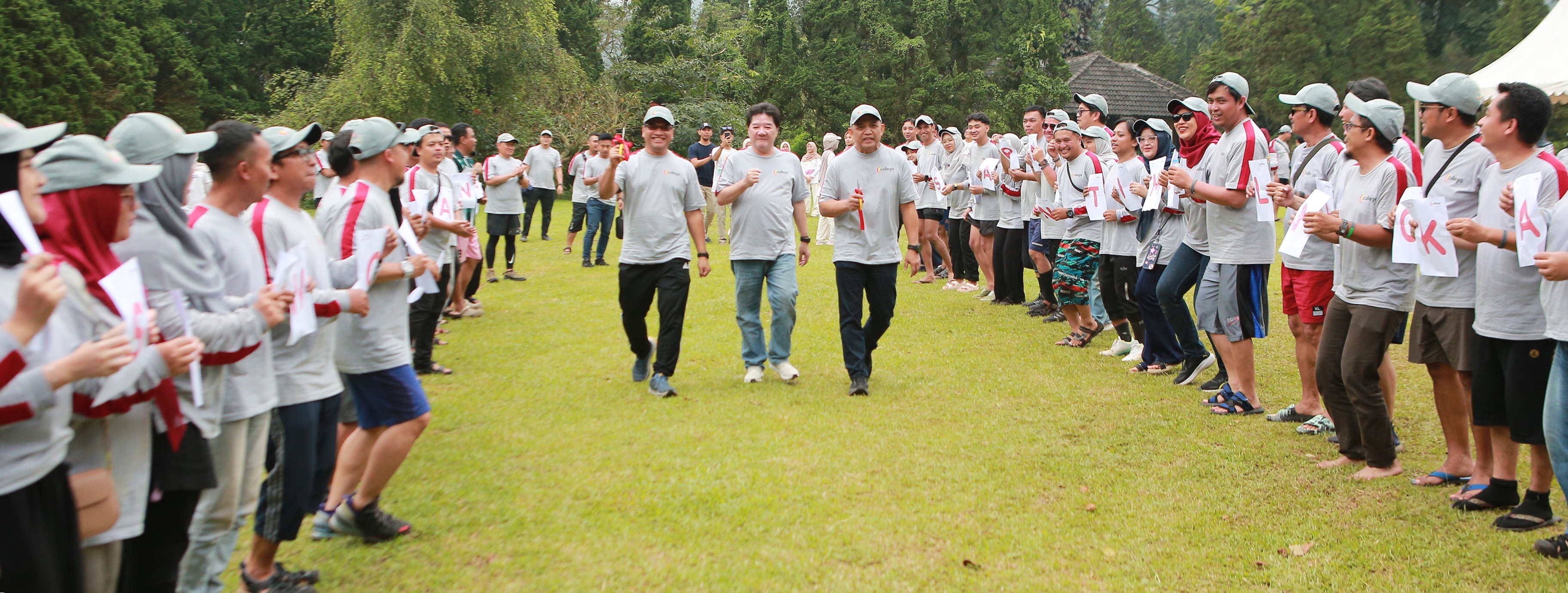Karyawan mengenakan kaos abu-abu dan topi berdiri berbaris di lapangan rumput membentuk lorong, menyemangati tiga pimpinan perusahaan yang berjalan di tengah dalam suasana outing dan team building di area terbuka dengan latar pepohonan.