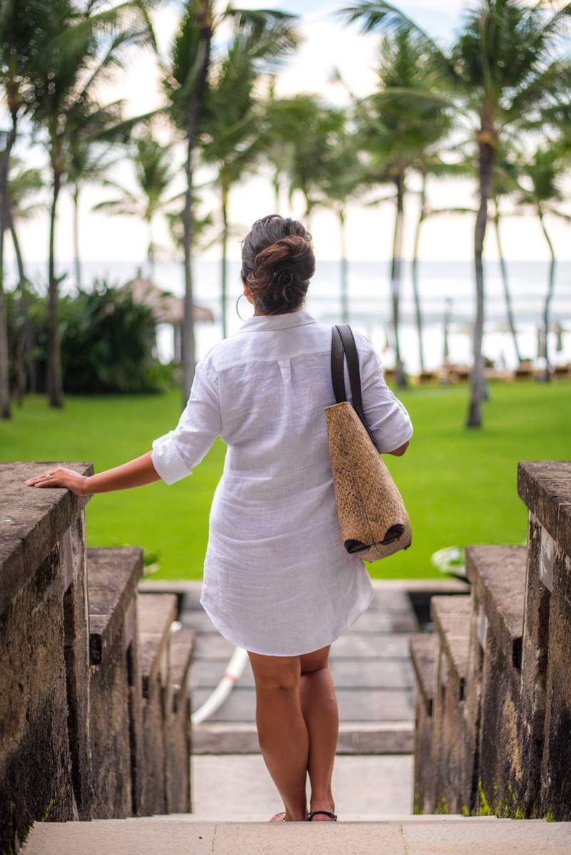 A woman in a white dress and a beach bag stands on a stone path leading toward a tropical beach with palm trees.