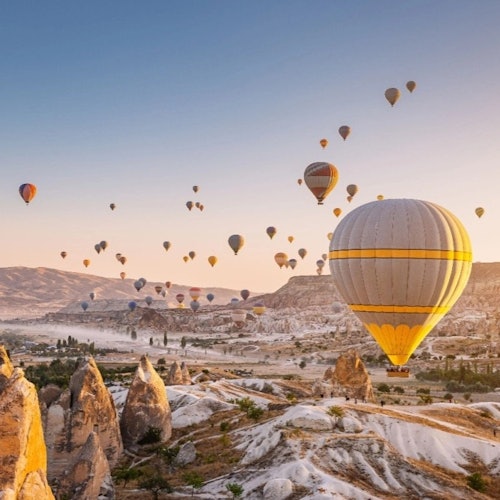 在日出时分,众多热气球漂浮在干旱岩石地貌的上空,背景中有山脉和山谷的映衬。