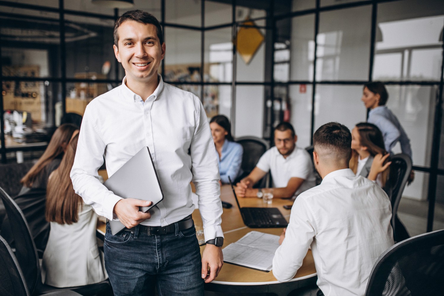 A man in a white shirt smiles while walking towards a meeting room with colleagues engaged in discussion at the table.
