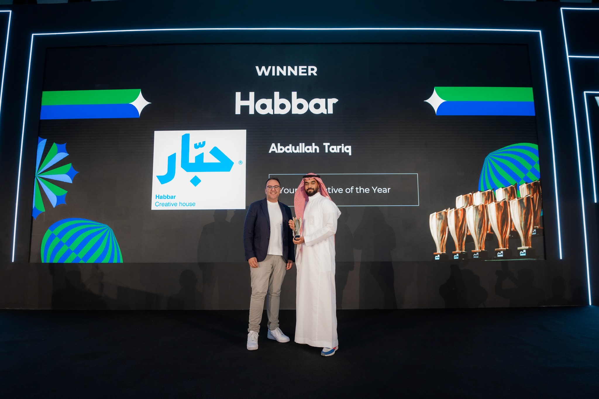 Three people in conversation with warm lighting, including a person wearing a hat and another person wrapped in what appears to be a textured shawl or sweater.