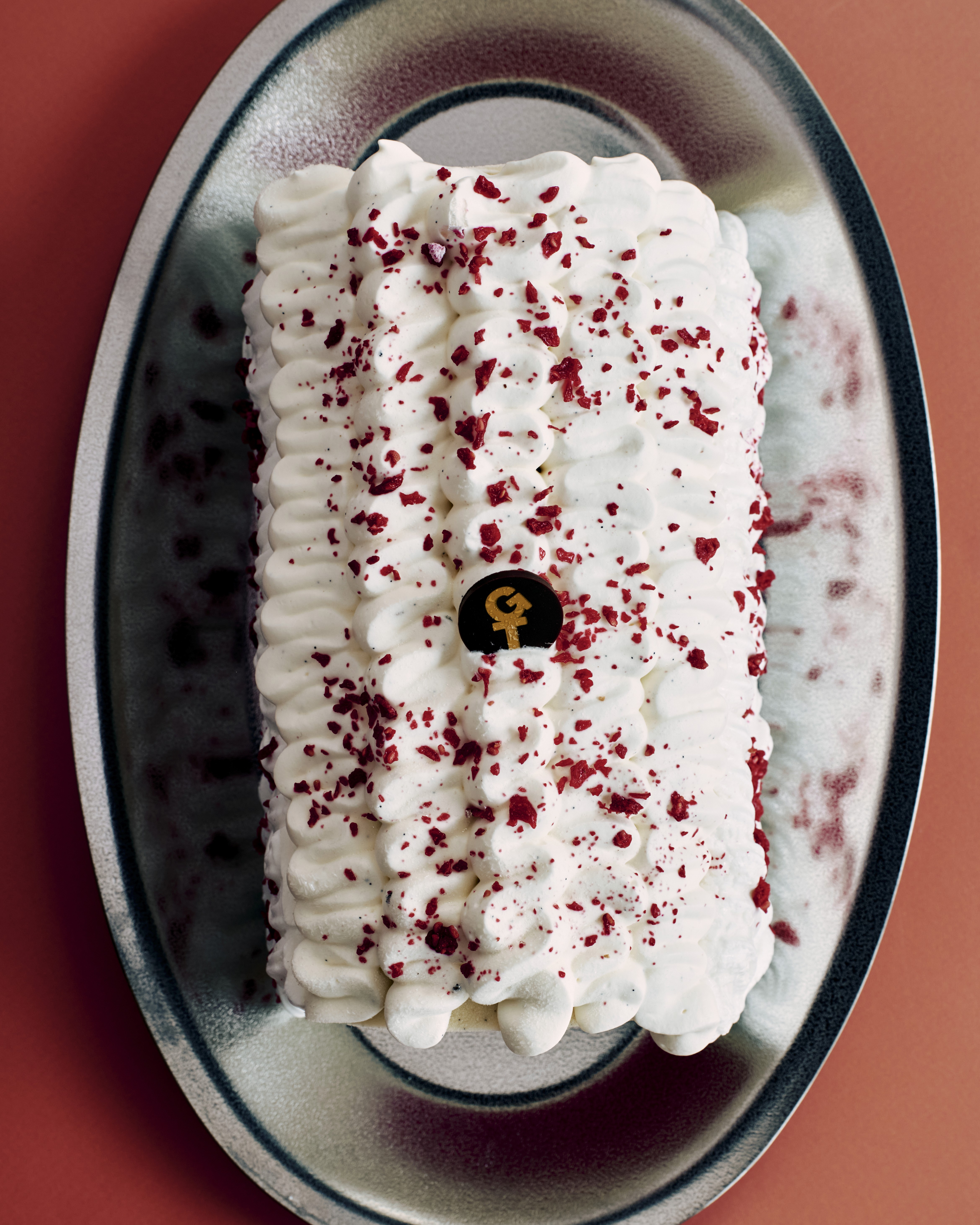 Gâteau rectangulaire recouvert de crème fouettée et de éclats rouges, avec logo 'GT' au centre